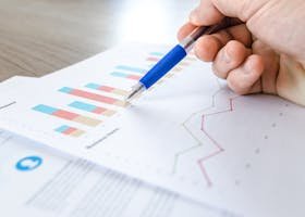 A close-up of a hand with a pen analyzing data on colorful bar and line charts on paper.
